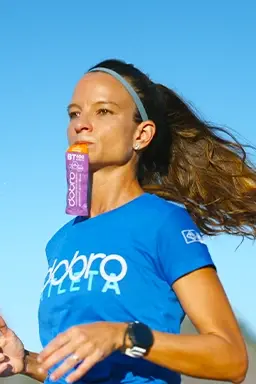 Mulher correndo ao ar livre com sachê Dobro BT sabor Pink Lemonade na boca, vestindo camiseta azul da equipe Dobro.