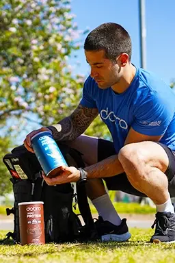 Atleta ajoelhado ao lado da mochila, segurando pote de suplemento Dobro ao ar livre. Outros produtos Dobro visíveis na grama.