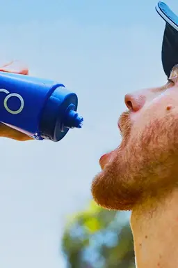 Homem barbudo bebendo de uma garrafa Dobro azul durante atividade ao ar livre em dia ensolarado.