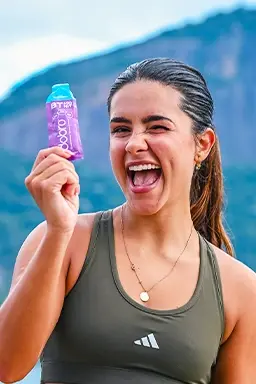 Mulher sorridente segurando sachê Dobro BT sabor Pink Lemonade, em cenário ao ar livre com montanhas ao fundo.