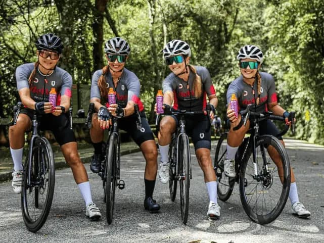 Quatro ciclistas mulheres uniformizadas, sorrindo e segurando sachês Dobro enquanto posam com suas bicicletas em uma estrada arborizada.